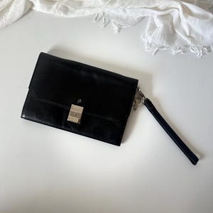 May include: Black leather clutch bag with a silver clasp and wrist strap. The rectangular bag features a flap closure and a minimalist design. A white scarf is in the background.