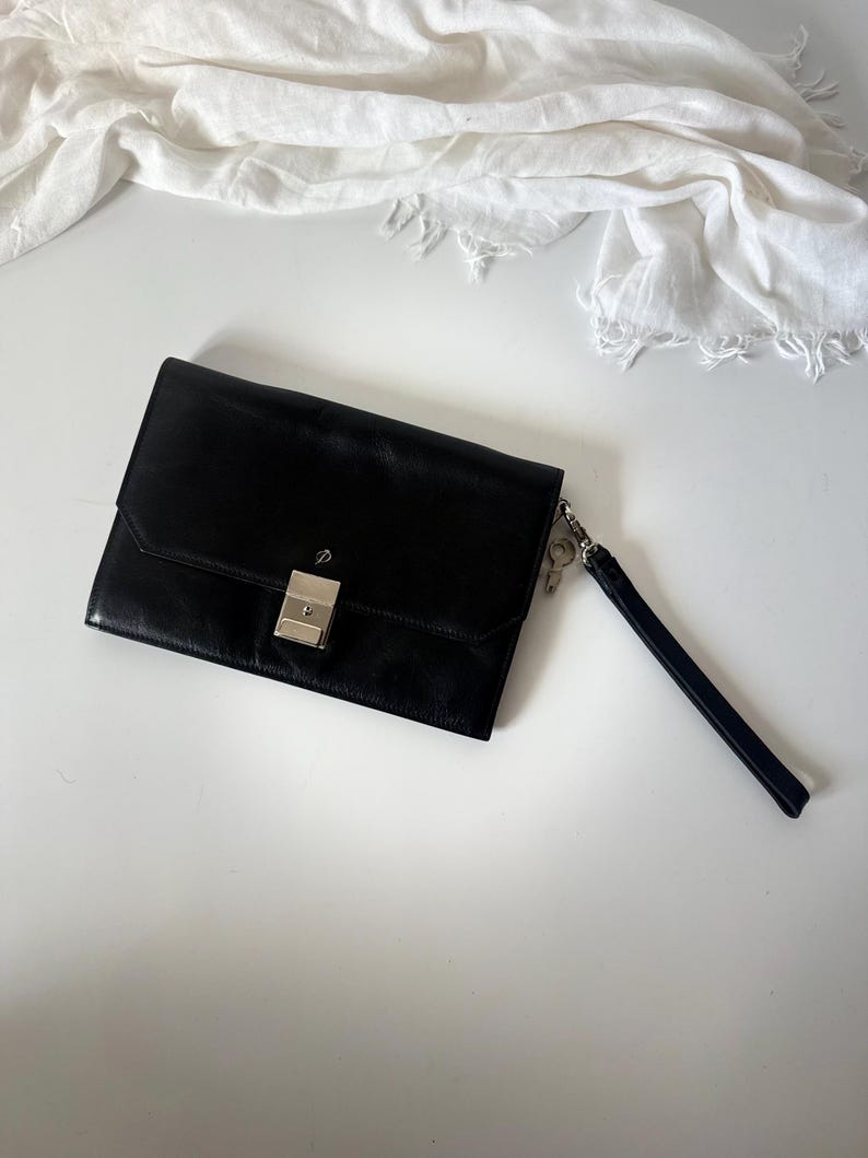 May include: Black leather clutch bag with a silver clasp and wrist strap. The rectangular bag features a flap closure and a minimalist design. A white scarf is in the background.