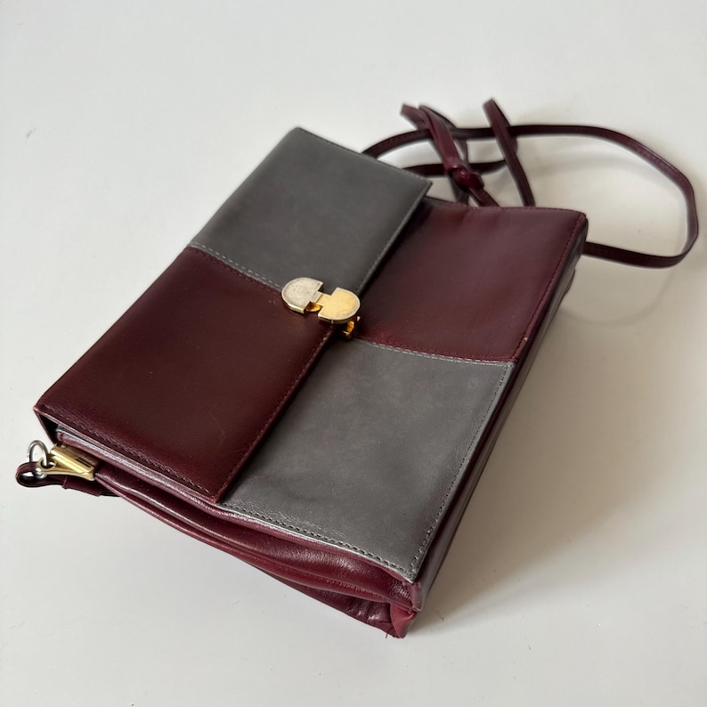 May include: A burgundy and gray leather shoulder bag with a gold-tone clasp. The bag features a rectangular shape with a flap closure and a long, thin strap. The bag is divided into color-blocked sections.