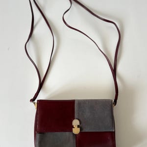 May include: A burgundy and gray crossbody bag with a long, thin strap. The bag is rectangular with a gold-colored clasp. The front of the bag is divided into two squares, one burgundy and one gray.