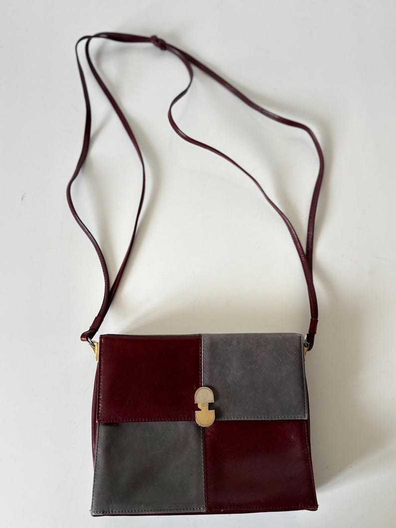 May include: A burgundy and gray crossbody bag with a long, thin strap. The bag is rectangular with a gold-colored clasp. The front of the bag is divided into two squares, one burgundy and one gray.