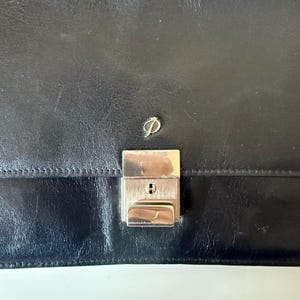 May include: A black leather briefcase with a silver metal clasp and a small silver logo. The briefcase is rectangular with a smooth, slightly textured surface. The clasp has a keyhole.