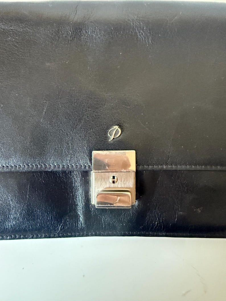 May include: A black leather briefcase with a silver metal clasp and a small silver logo. The briefcase is rectangular with a smooth, slightly textured surface. The clasp has a keyhole.