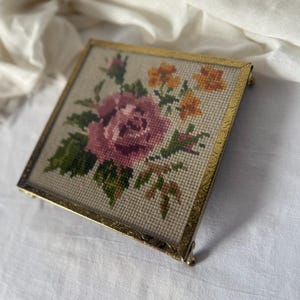 May include: A square, vintage needlepoint picture with a gold-colored metal frame. The needlepoint features a large pink rose with green leaves and smaller orange flowers. The frame has decorative detailing and small feet.