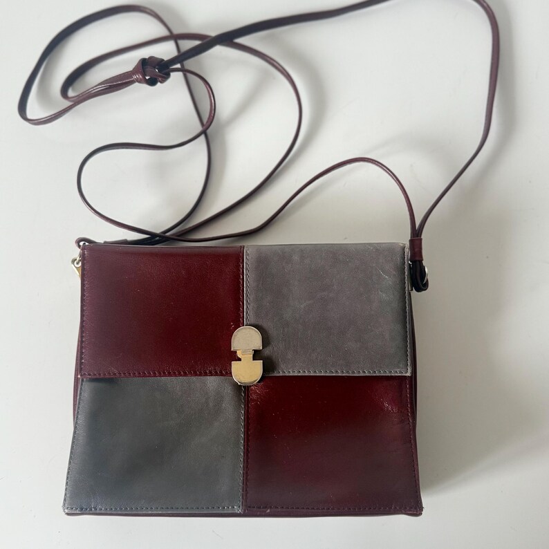 May include: A burgundy and gray leather crossbody bag with a long, thin strap. The bag is divided into four sections, two burgundy and two gray, with a gold-colored clasp.