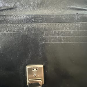 May include: Black leather wallet with a silver clasp. The interior has multiple card slots and a small embossed logo. The wallet is closed, showing the clasp and the smooth leather exterior.