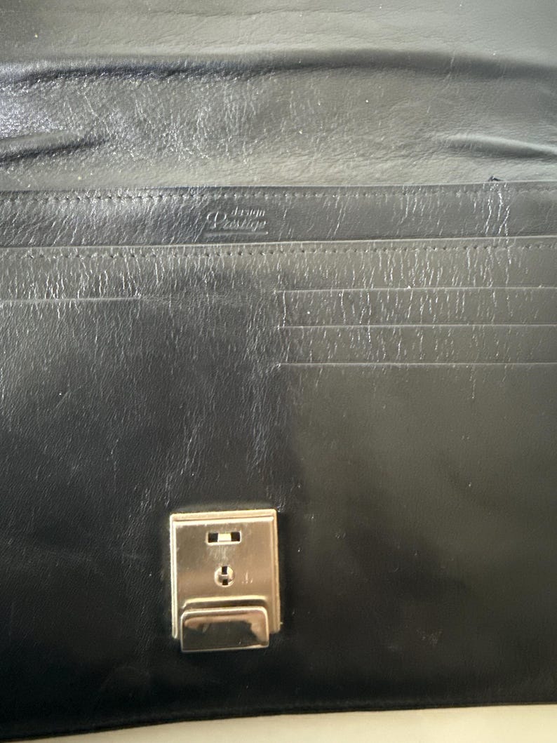 May include: Black leather wallet with a silver clasp. The interior has multiple card slots and a small embossed logo. The wallet is closed, showing the clasp and the smooth leather exterior.