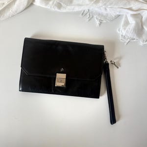 May include: A black leather clutch bag with a silver clasp and a wrist strap. The rectangular bag has a small key attached to the strap. A white fabric is in the background.