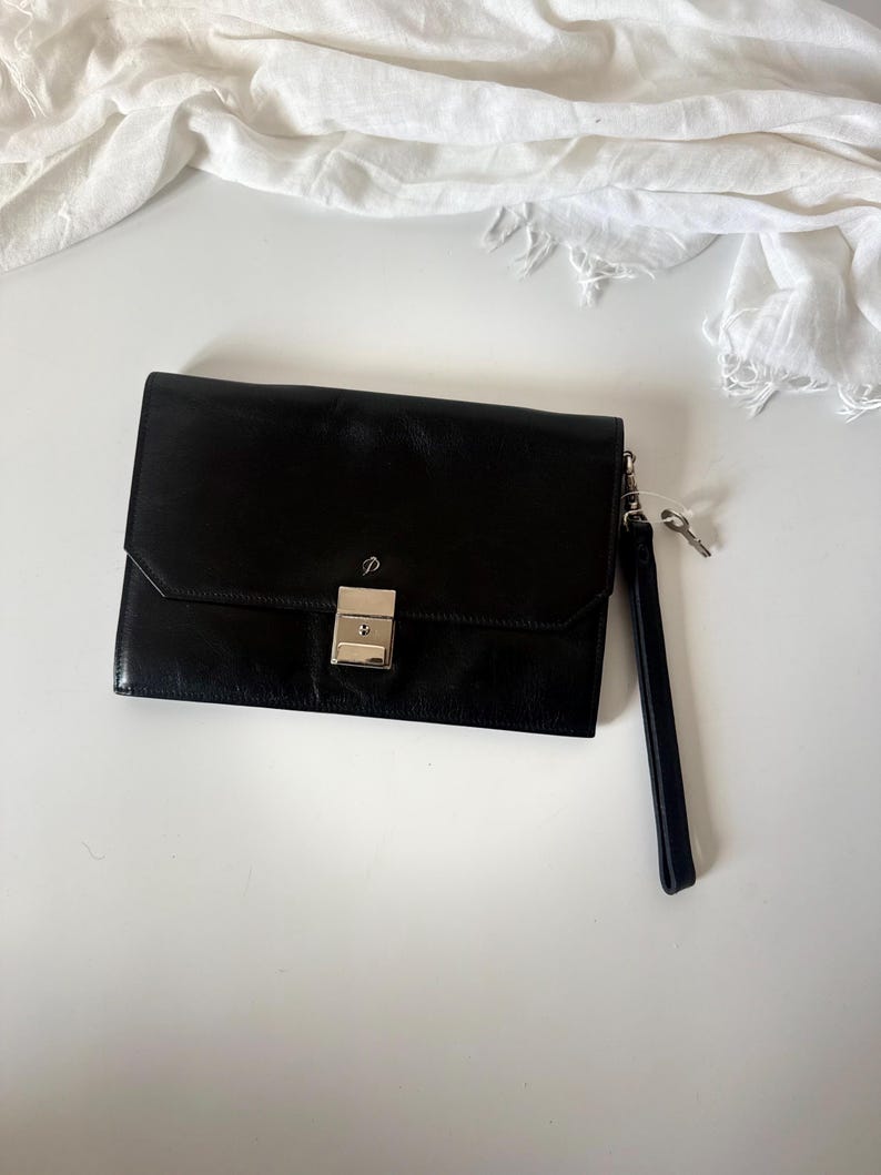 May include: A black leather clutch bag with a silver clasp and a wrist strap. The rectangular bag has a small key attached to the strap. A white fabric is in the background.