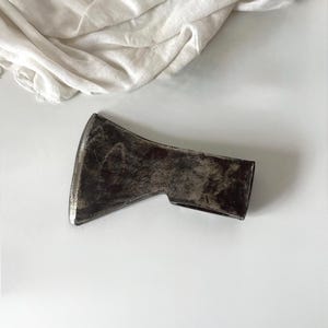 May include: A vintage axe head with a weathered, metallic finish. The axe head has a dark, aged appearance, with visible signs of wear and use. The blade is sharp and ready for use. A white cloth is in the background.