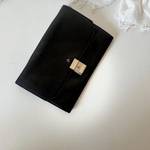 May include: A black leather clutch bag with a silver clasp. The rectangular bag is closed and has a flap closure. The clutch is on a white surface with a white cloth in the background.