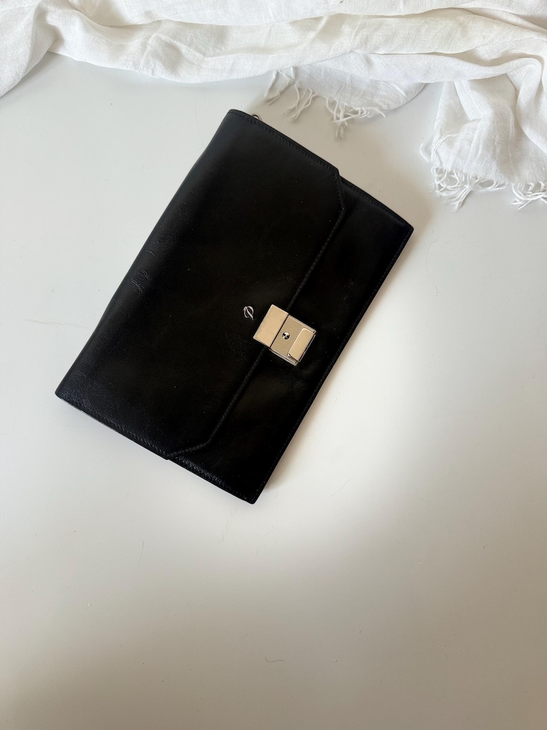 May include: A black leather clutch bag with a silver clasp. The rectangular bag is closed and has a flap closure. The clutch is on a white surface with a white cloth in the background.