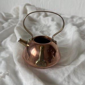 Könnte beinhalten: Eine kleine, kupferfarbene Teekanne mit einem silbernen Griff und Ausguss. Die Teekanne hat ein Messingemblem auf der Vorderseite. Die Teekanne steht auf einer weißen, strukturierten Oberfläche.