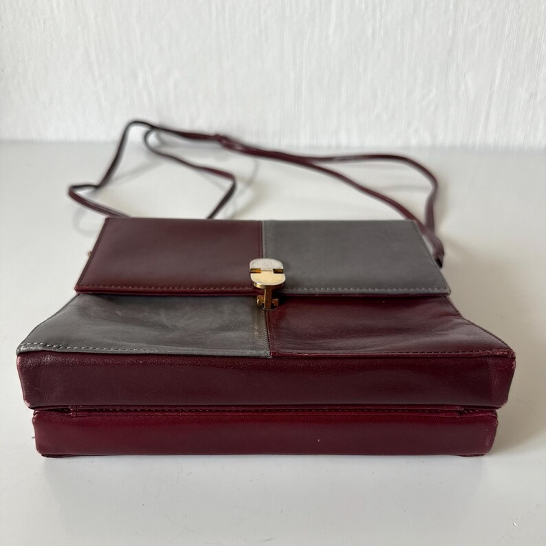 May include: A two-tone crossbody bag in burgundy and gray leather. The bag features a rectangular shape with a flap closure secured by a gold-tone clasp. A thin, burgundy strap extends from the bag for shoulder or crossbody wear.