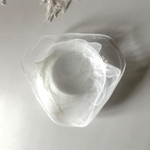 Op de afbeelding: Een heldere, witte glazen kom met een unieke, onregelmatige vorm. De kom heeft een getextureerd, gemarmerd uiterlijk, met wervelende witte patronen door het glas. De kom wordt waarschijnlijk gebruikt voor decoratieve doeleinden of om te serveren.