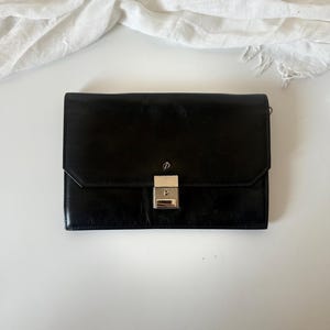 May include: A black leather clutch bag with a silver clasp. The rectangular bag has a flap closure and a small, silver emblem above the clasp. A white cloth is in the background.
