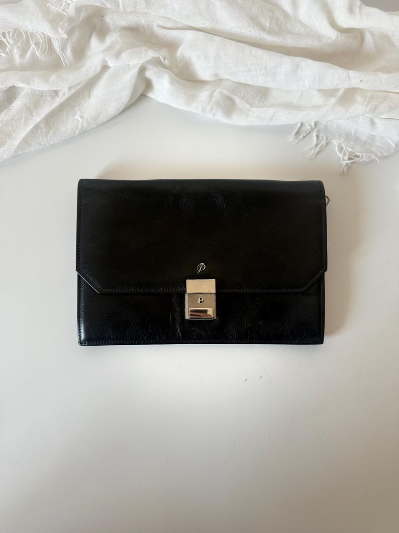 May include: A black leather clutch bag with a silver clasp. The rectangular bag has a flap closure and a small, silver emblem above the clasp. A white cloth is in the background.