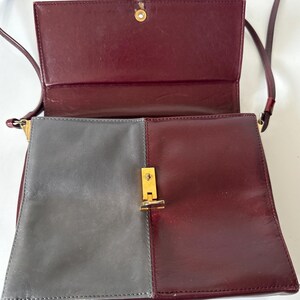May include: A two-tone shoulder bag with a flap closure. The bag features a color block design with a gray panel on the left and a burgundy panel on the right. The flap and strap are burgundy. The bag has a gold-colored clasp.