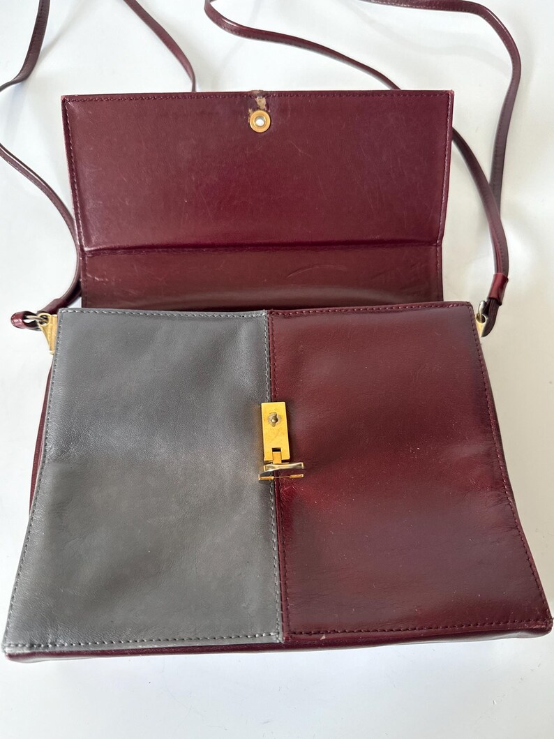 May include: A two-tone shoulder bag with a flap closure. The bag features a color block design with a gray panel on the left and a burgundy panel on the right. The flap and strap are burgundy. The bag has a gold-colored clasp.