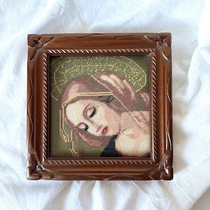 Può includere: Ritratto a punto croce incorniciato di una donna con capelli castani e aureola dorata. L'opera è ambientata in una cornice quadrata di legno marrone ornato. Lo sfondo è verde tenue.