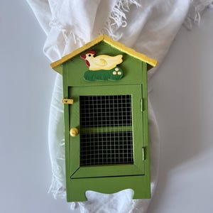 May include: A green wooden chicken coop with a yellow roof and a chicken design. The coop has a wire mesh door and a small yellow knob. The chicken is yellow with a red comb and is perched on a patch of green grass.