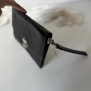 May include: Black leather clutch bag with a silver clasp and a wrist strap. The rectangular bag has a flap closure and a small key attached. The clutch is on a white surface.