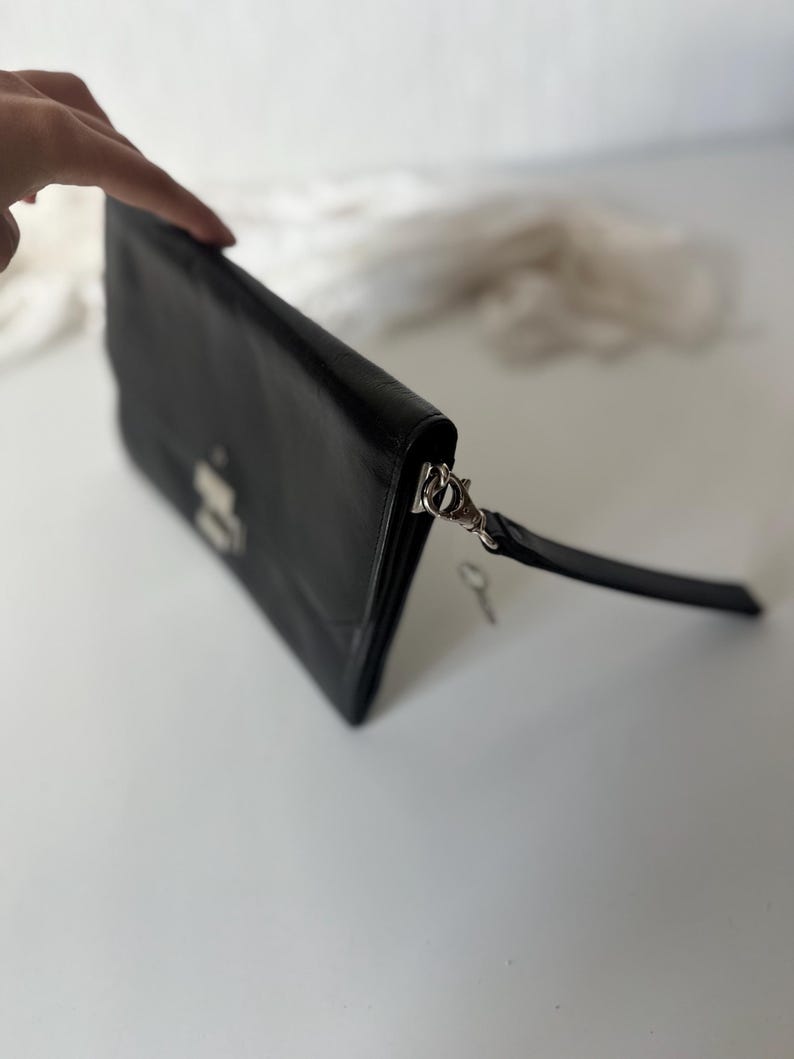 May include: Black leather clutch bag with a silver clasp and a wrist strap. The rectangular bag has a flap closure and a small key attached. The clutch is on a white surface.