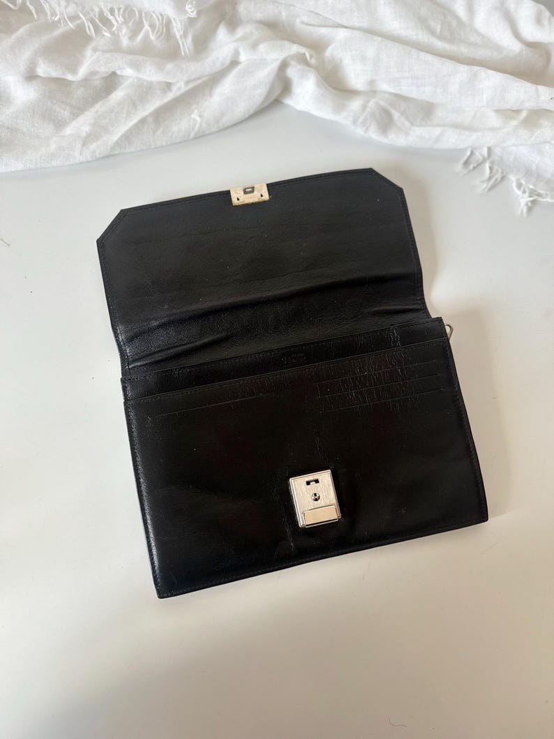 May include: Black leather wallet with a silver clasp. The wallet is open, showing several card slots. The rectangular wallet has a shiny finish and a classic design. The background is white.