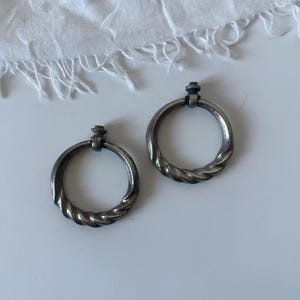 May include: A pair of silver-coloured hoop earrings featuring a twisted rope pattern. Each earring has a small decorative top. The earrings are presented on a white surface with a white, fringed fabric in the background. These earrings are a classic jewellery item.
