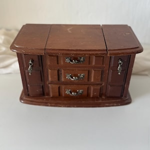 Op de afbeelding: Een vintage houten sieradendoosje met een donkerbruine afwerking. De doos heeft een rechthoekige vorm met een scharnierend deksel en twee zijcompartimenten. Het heeft drie kleine lades met sierlijke metalen handgrepen en decoratieve accenten.