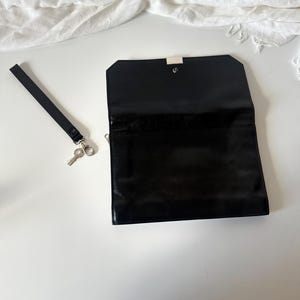 May include: Black rectangular clutch with a silver clasp, open to reveal the interior. A black wrist strap with a silver clasp and a small key are to the left of the clutch. The clutch is on a white surface.