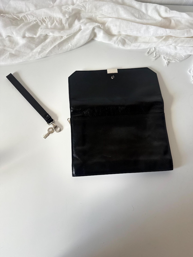 May include: Black rectangular clutch with a silver clasp, open to reveal the interior. A black wrist strap with a silver clasp and a small key are to the left of the clutch. The clutch is on a white surface.