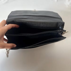 May include: Black leather shoulder bag with a silver clasp. The bag is open, revealing a zippered pocket and a spacious interior. The bag's design is simple and classic, suitable for everyday use. The bag is made of a smooth, matte leather.