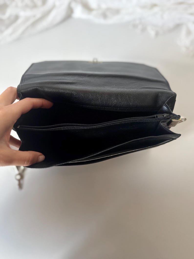 May include: Black leather shoulder bag with a silver clasp. The bag is open, revealing a zippered pocket and a spacious interior. The bag's design is simple and classic, suitable for everyday use. The bag is made of a smooth, matte leather.