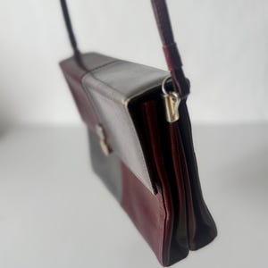 May include: A rectangular crossbody bag in a color block design. The bag features a flap closure and a long, thin strap. The color scheme includes burgundy, gray, and a darker shade of burgundy. The bag is made of a smooth material.