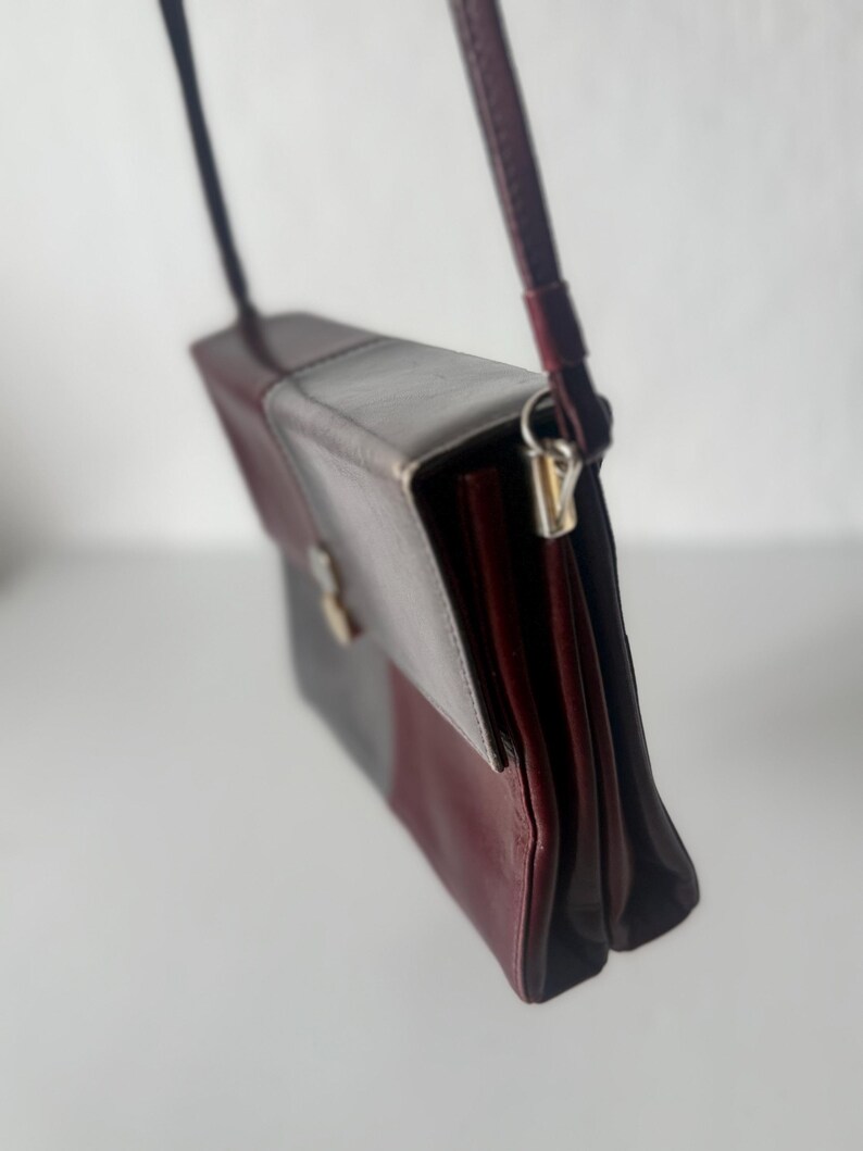 May include: A rectangular crossbody bag in a color block design. The bag features a flap closure and a long, thin strap. The color scheme includes burgundy, gray, and a darker shade of burgundy. The bag is made of a smooth material.