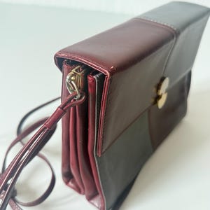 May include: A burgundy and gray leather crossbody bag with a long, thin strap. The bag has a rectangular shape with a flap closure and a gold-colored clasp. The bag is made of leather and has a stitched design.