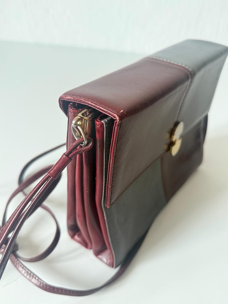 May include: A burgundy and gray leather crossbody bag with a long, thin strap. The bag has a rectangular shape with a flap closure and a gold-colored clasp. The bag is made of leather and has a stitched design.