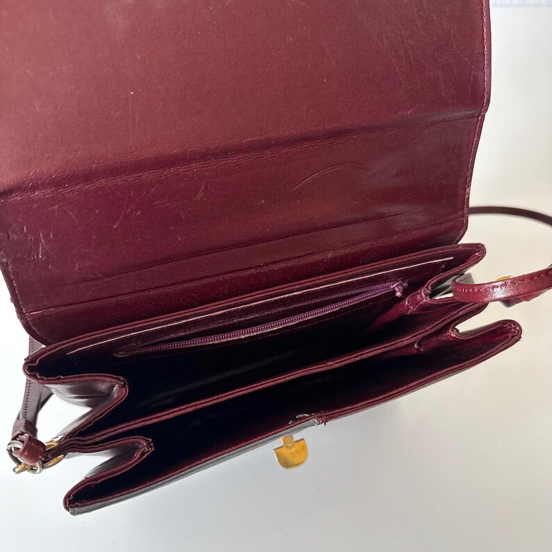 May include: Burgundy leather crossbody bag with a gold-tone clasp. The bag has multiple compartments and a long, thin strap. The interior features a zippered pocket. The bag is open, revealing the inside.