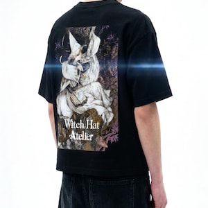 May include: A black short-sleeve t-shirt featuring a large rectangular graphic on the back. The print depicts a character in a pointed hat and flowing robes, with the text Witch Hat Atelier printed in white below the illustration.