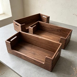 Può includere: Tre vassoi rettangolari in legno marrone scuro con venature visibili. Ogni vassoio presenta un'apertura frontale ribassata, maniglie laterali integrate e angoli con giunzioni a pettine, offrendo un design aperto per organizzare oggetti.