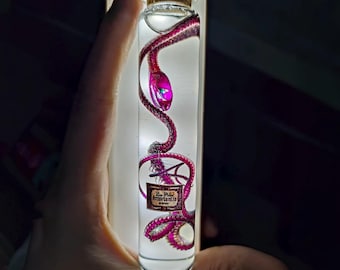 Op de afbeelding: Een heldere glazen fles met een kurk, met daarin een roze en paarse slangenbeeld. De slang is opgerold in de fles, met een label met de tekst "Amortentia". De fles wordt tegen een helder licht gehouden.