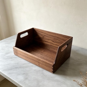 May include: A rectangular wooden storage tray crafted from dark brown walnut wood with a visible grain pattern. The tray features high side walls, a lower front panel, and integrated cutout handles on both sides with box joint construction.