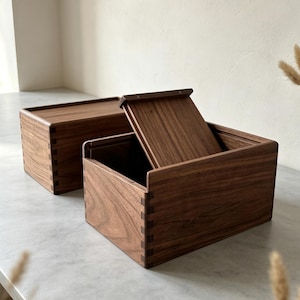 May include: Two rectangular dark brown wooden boxes with visible grain patterns and finger joint construction. One box is closed with a flat lid, while the other box is open with its matching wooden lid resting at an angle against the back edge.
