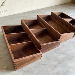 Può includere: Un set di tre vassoi rettangolari in legno marrone scuro con venature visibili. Ogni vassoio presenta angoli con giunzioni a pettine e un divisorio interno, creando due scomparti separati all'interno di ciascun contenitore in legno.