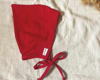 Könnte beinhalten: Eine einfarbige rote Haube mit abgerundeter Form und langen Stoffbändern zum Binden unter dem Kinn. Ein kleines weißes rechteckiges Etikett mit dem schwarzen Text Leo & Lola ist an der Seite der Haube nahe der Bänder angenäht.