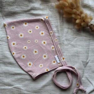 May include: A lilac baby bonnet featuring a daisy print. The bonnet has a tie fastening and a small label that reads "Leo & Lola". The daisies have white petals and yellow centres. The bonnet is made of soft material.
