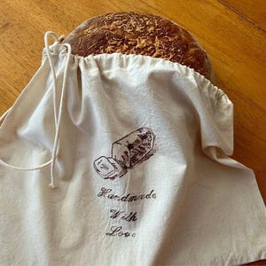 May include: A white cotton bread bag with a brown drawstring and a brown graphic of a loaf of bread with the text "Handmade with Love".