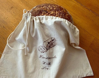 May include: A white cotton bread bag with a brown drawstring and a brown graphic of a loaf of bread with the text "Handmade with Love".