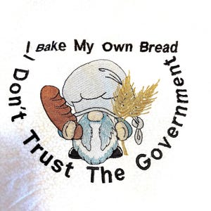 Puede incluir: Una toalla de cocina blanca bordada con un gnomo que lleva un gorro de cocinero y sostiene una hogaza de pan y una espiga de trigo. El texto alrededor del gnomo dice "No confíes en el gobierno, yo horneo mi propio pan".